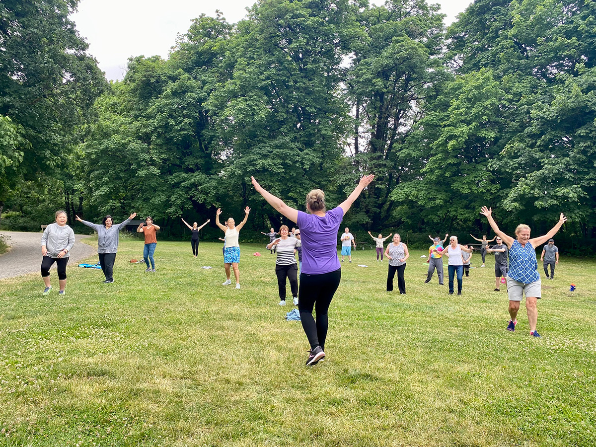 Community members take a yoga class outdoors in a grassy clearing near tall trees
