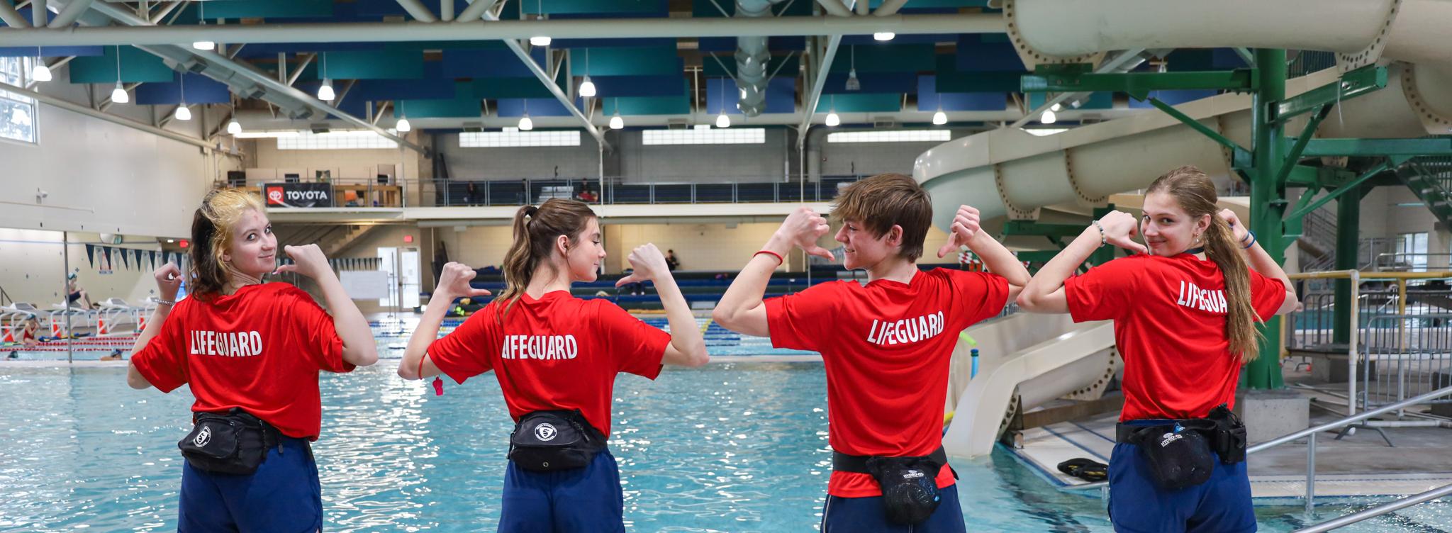 Four young lifeguards point to their lifeguard shirts while posing in front of the pool
