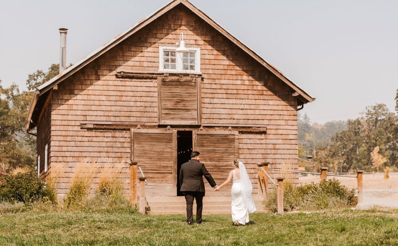 A bride and groom hold hands as they enter a barn from a grassy meadow