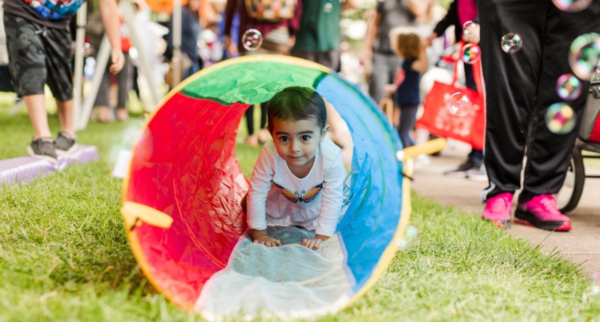 A child crawls through a rainbow-colored tube laying on top of green summer grass.