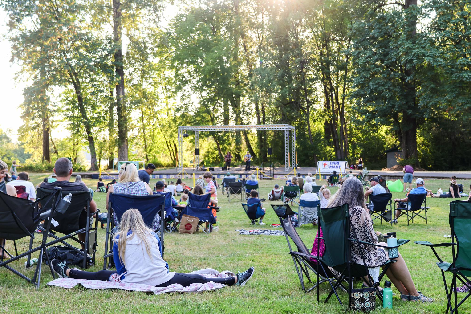 a crowd sits in lawn chairs at Island Park watching a concert on the stage