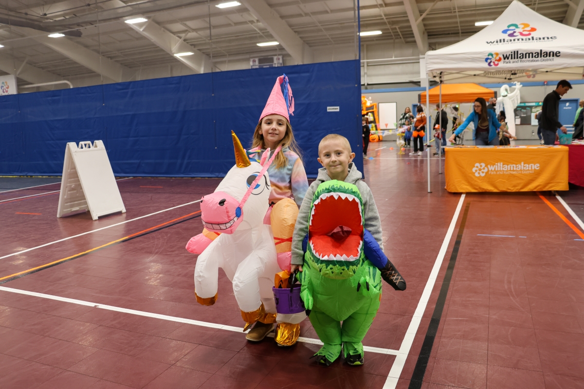 Two children posing in their halloween costumes in a gym. They are dressed as a unicorn and a dinosaur.