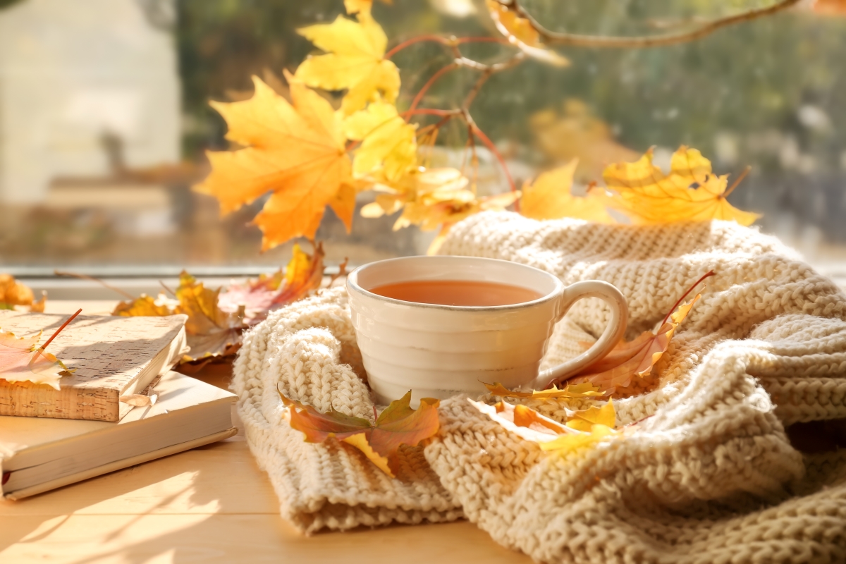 A cup of tea sits steaming in front of a background of fall leaves and a cozy sweater.