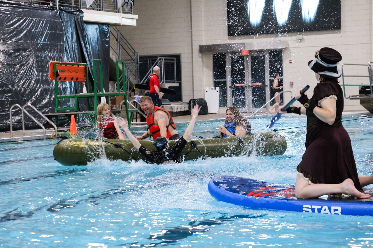 Three people sit in a green raft floating in a pool, while someone pops up from underneath the water's surface next to them.