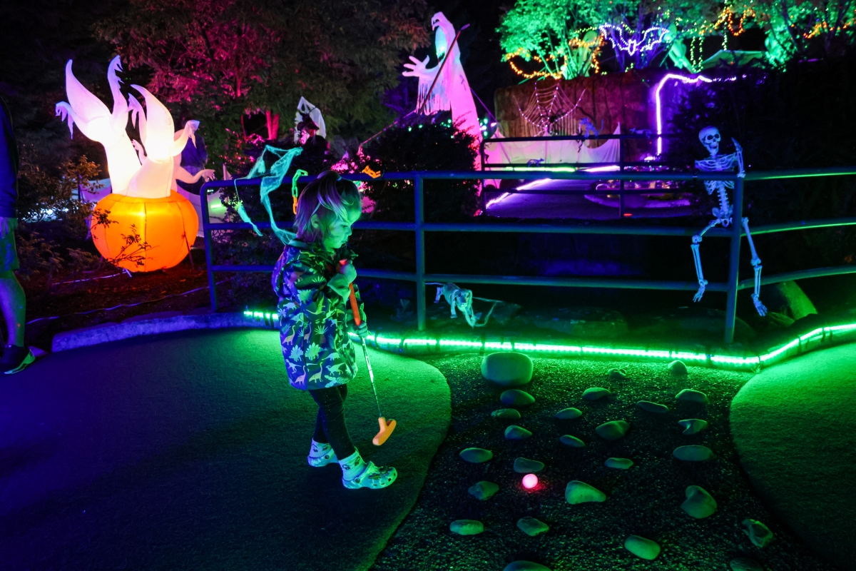 A girl stands on a dark mini golf course with a glowing pink ball and lighted Halloween decorations in the background.