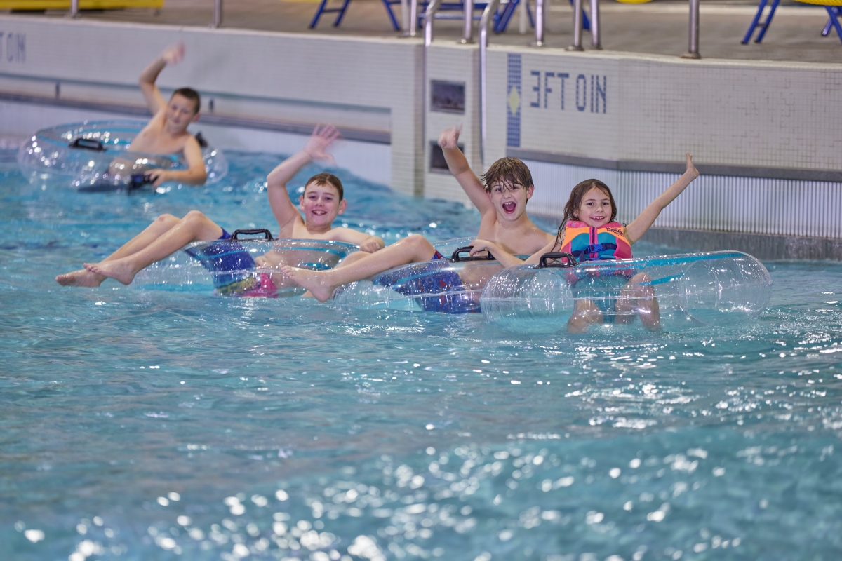 Four kids float on pool water while sitting on top of inflatable tubes. They are smiling and waving.