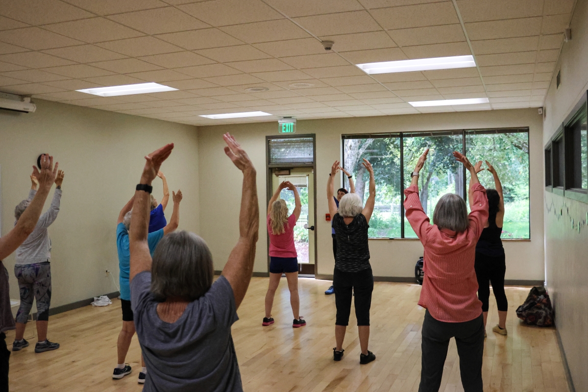 Adults stand with their hands over their heads in a yoga stretching pose in an indoor room.