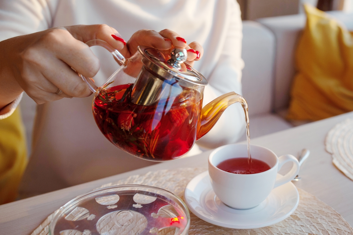 A clear tea pot with dark-colored tea, pouring into a white teacup.