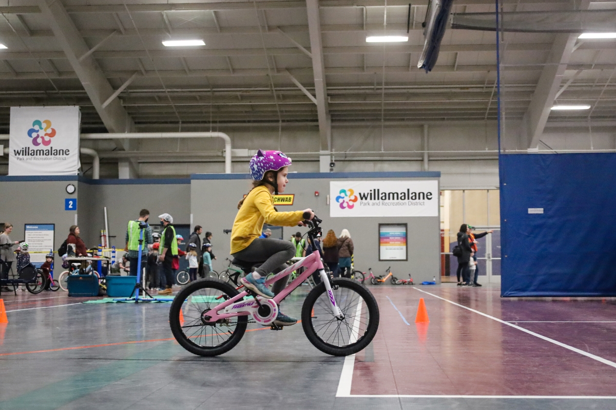 A child with a yellow shirt and purple bike helmet rides a small bike through a gym.