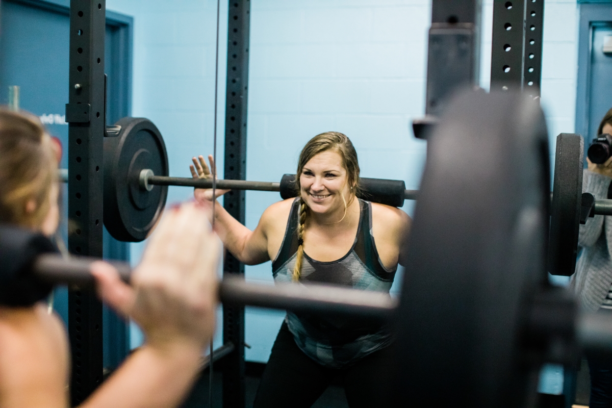 An adult smiling while lifting a weight bar over their shoulders.