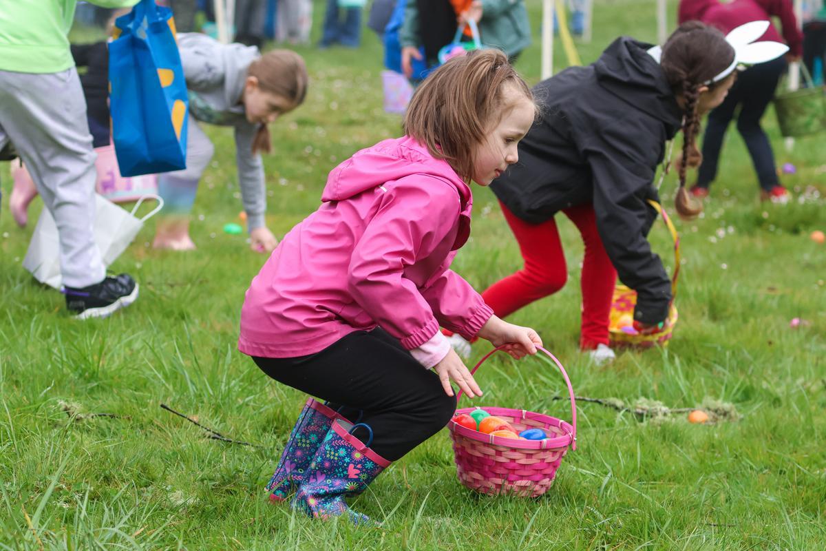 A young child in a pink coat holds a basket filled with colored eggs, while leaning down to pick up another in the green grass.