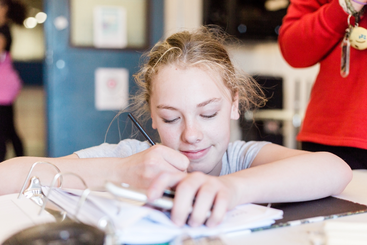 A teenager sits at a desk, writing on a piece of paper in a binder.