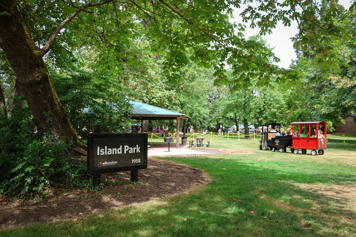 A red kid's train drives on the lawn behind a park sign that says "Island Park"