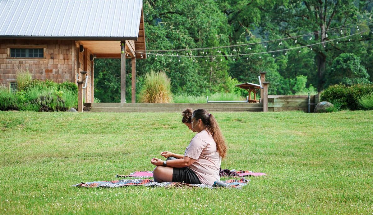 Two adults sit on a yoga mat outdoors on a grassy field.