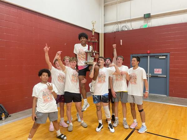 nine men pose in the gym wearing matching white shirts and hoisting up one player who is holding a giant trophy