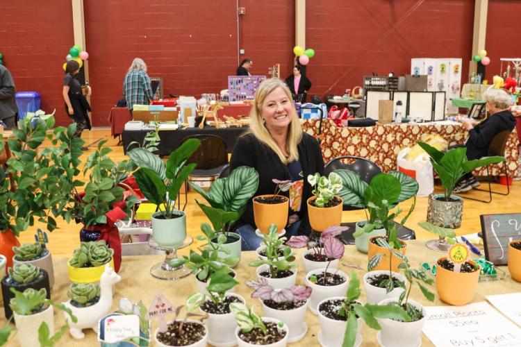 Lady sits at a table behind a bunch of houseplants at marketplace