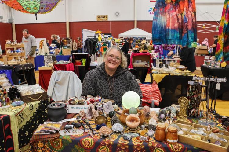 A vendor in an indoor marketplace sits behind a table of crystal goods