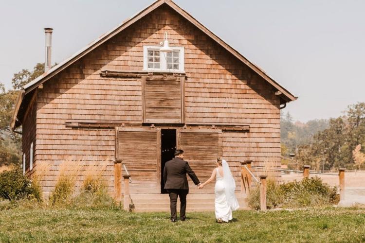 A bride and groom hold hands as they enter a barn from a grassy meadow