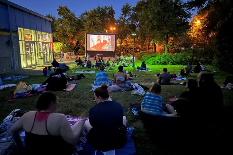 A group of people sit on the grass, enjoying a movie screened on a projector screen.