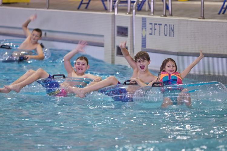 Four kids float on pool water while sitting on top of inflatable tubes. They are smiling and waving.