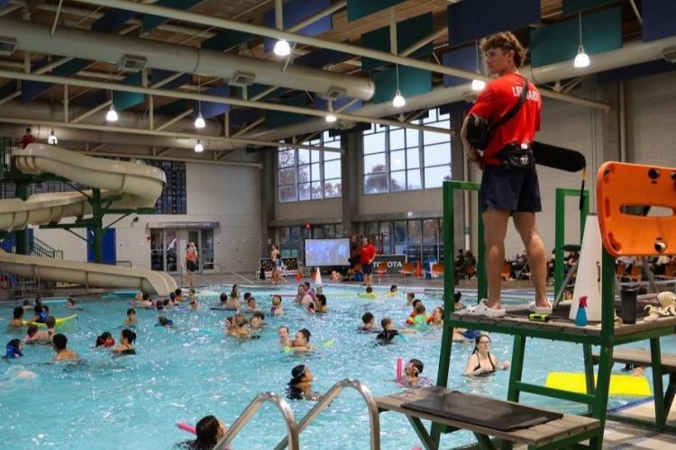 A lifeguard stands high next to a pool filled with swimmers.