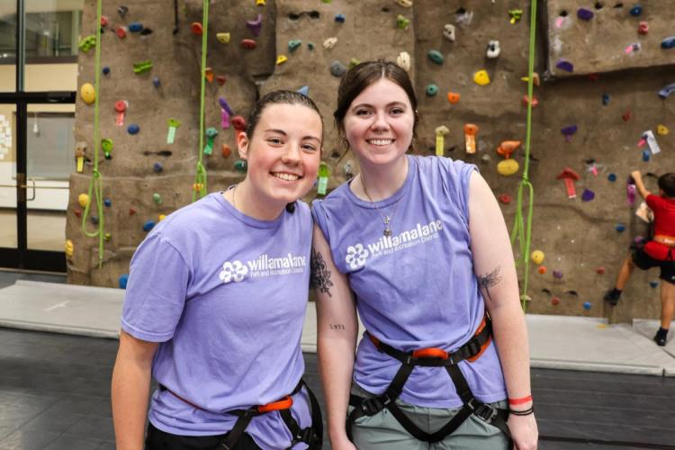 Two adults stand in front of a climbing wall in full gear.