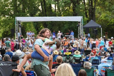 An adult and child dance together in the grass of an outdoor concert.
