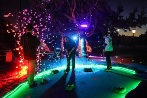 Three mini golfers stand on the green at night, with colorful lights and bubbles in the background.