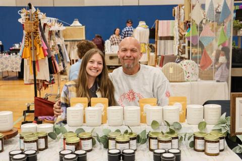 Two adults sit behind a table with plants for sale.