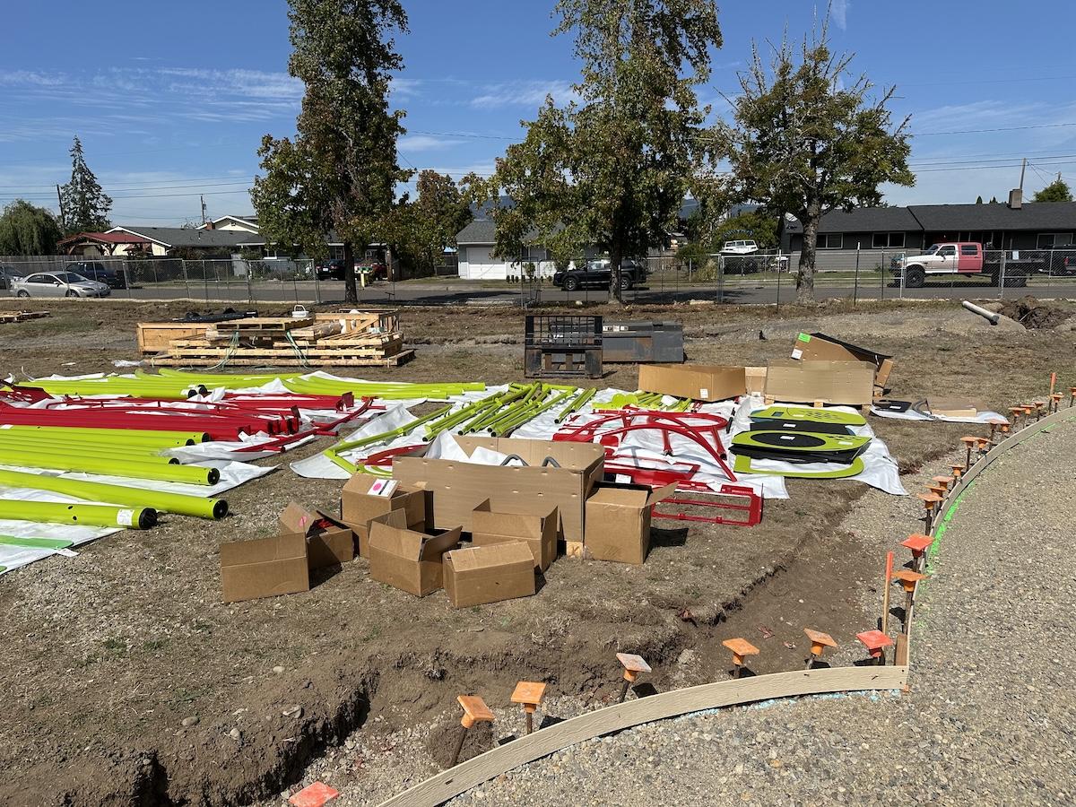 Colored playground equipment pieces lay in stacks on a dirt area where the new playground will go, once constructed.