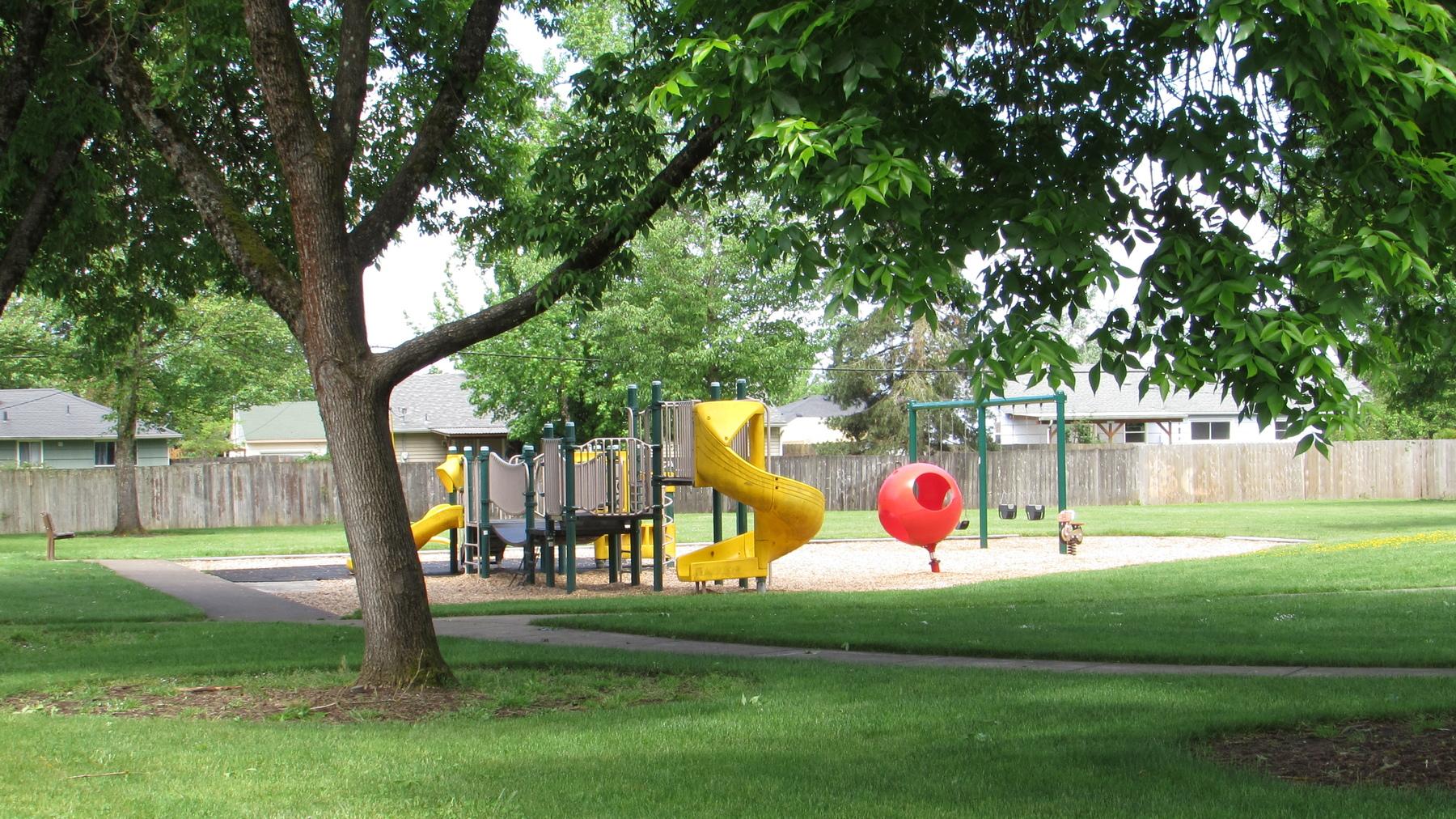 A tree and paved walking path lead to a small play structure with a slide and swings