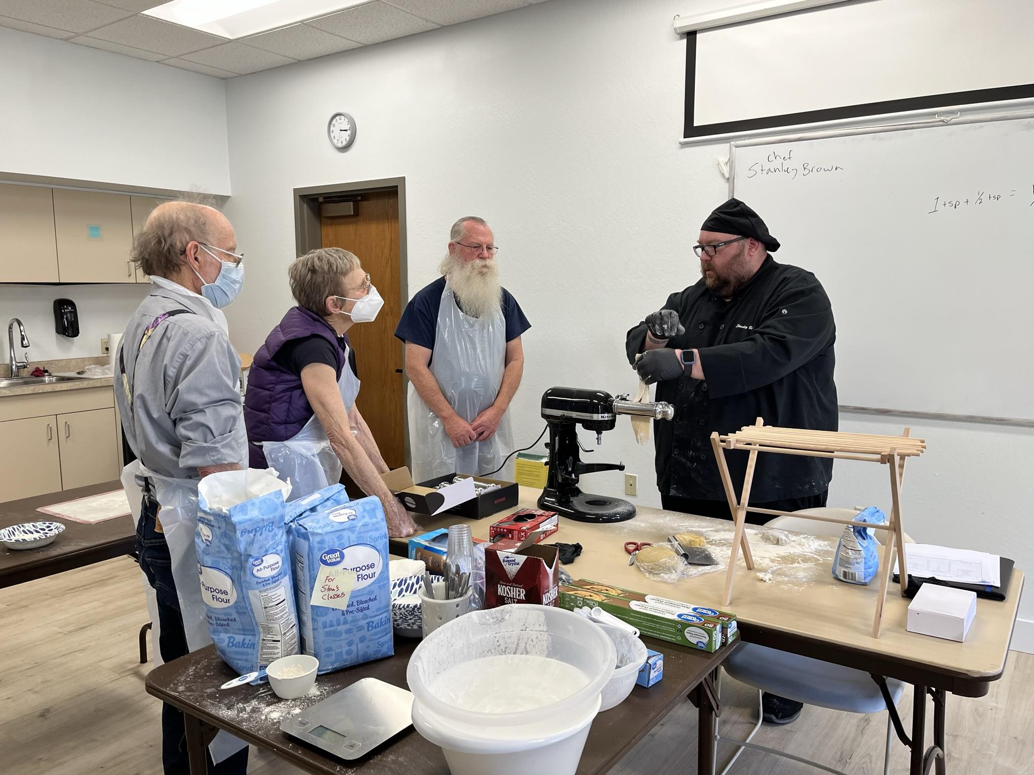Chef demonstrates pasta technique to a group of adult onlookers.
