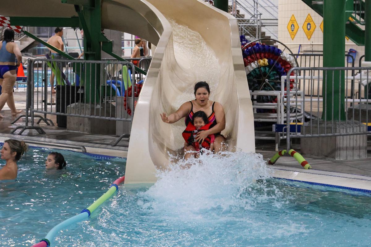 An adult and child ride down a water slide, about to splash into a pool.