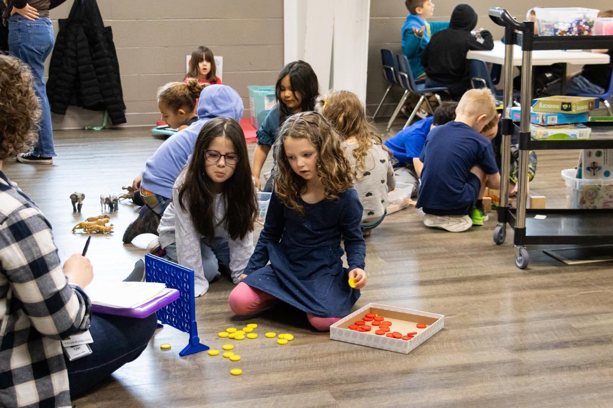Two kids sit in front of a connect four game, talking strategy.