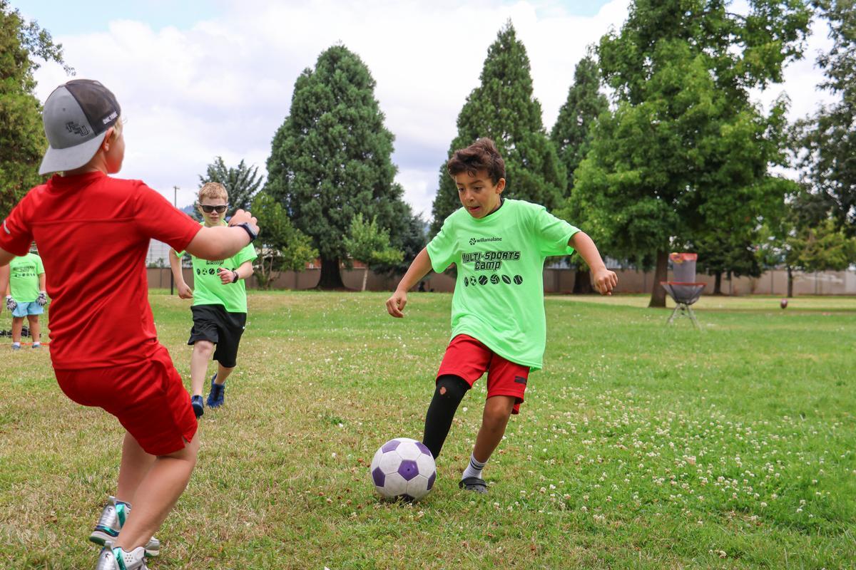 A child in a green shirt dribbles a soccer ball on a grassy field.