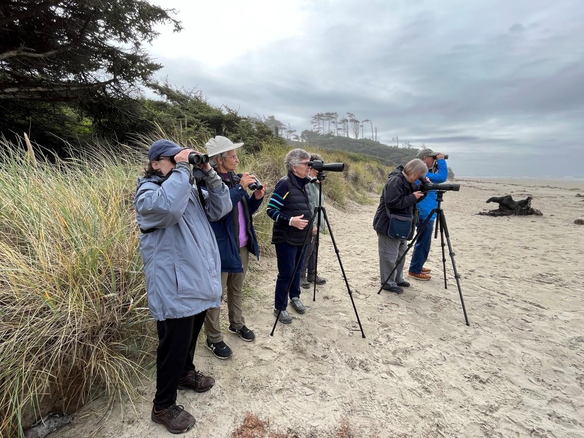 Five adults in coats standing on sand hold up binoculars, and look at nature from a distance.