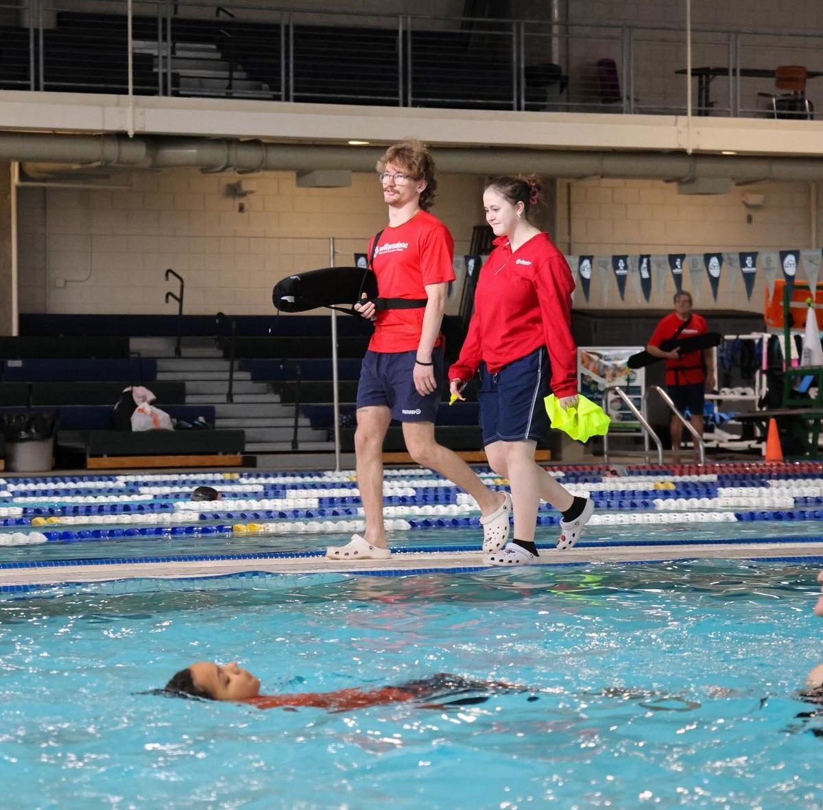 Two lifeguards in red shirts walk side-by-side on the pool's edge.