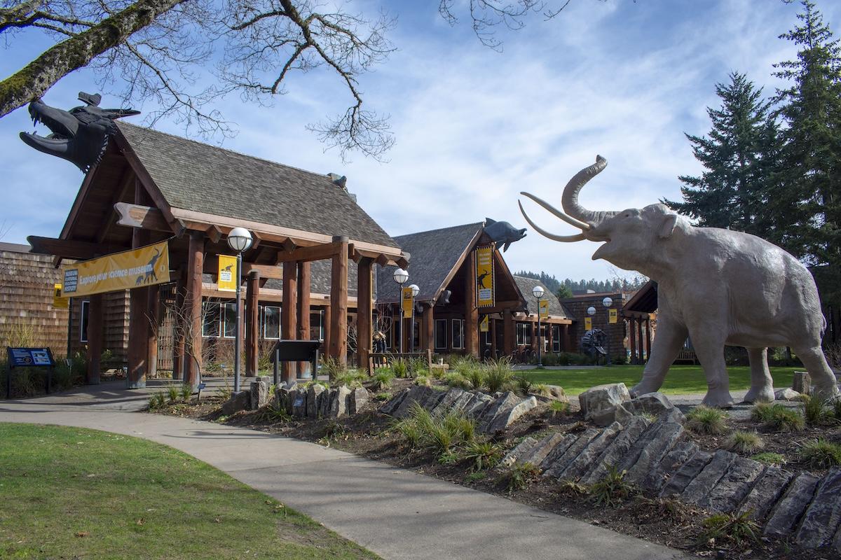 A mammoth statue in a grass field in front of a log cabin-style building.