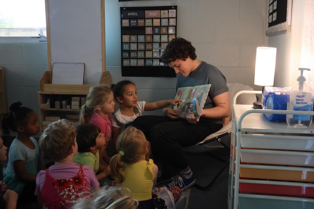 A group of children sit around an adult reading a book, with one pointing at it.