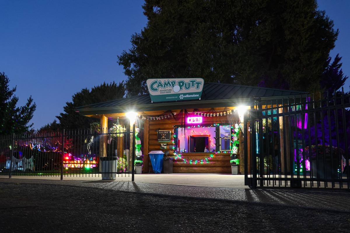 A building behind a gate at dusk, with two bright lights and colorful ones shining in the distance.