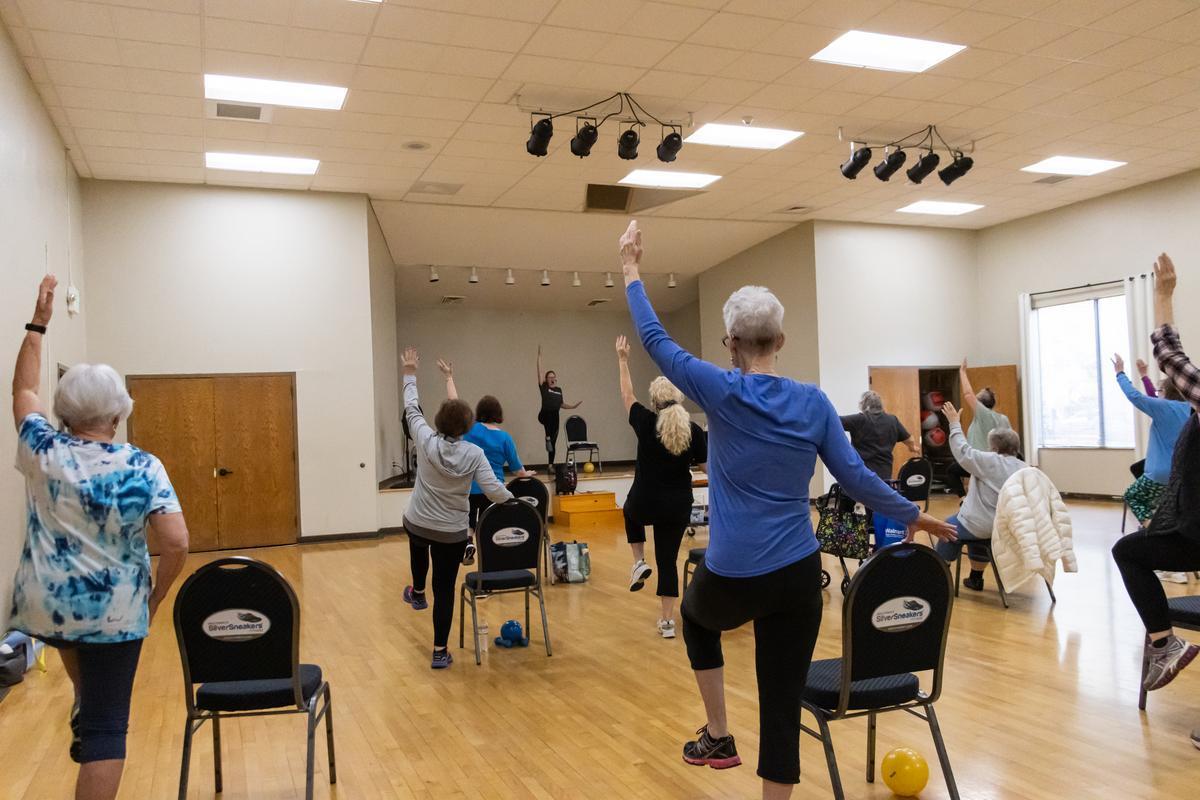 A group of adults holding chairs with one hand and the other in the air in a wood gym.