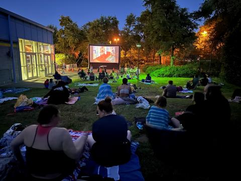 A group of people sit on the grass, enjoying a movie screened on a projector screen.