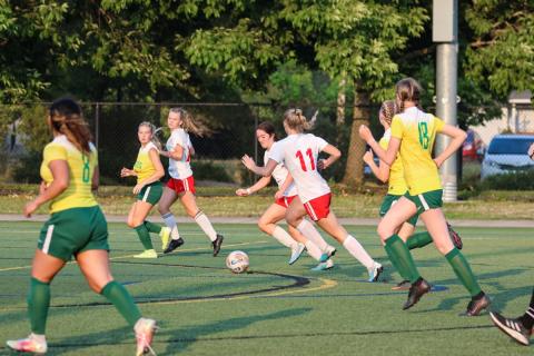 A group of soccer players chasing the ball during a competitive game on a green turf field.