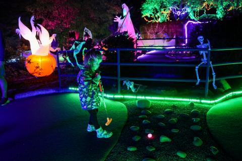 A child holds a golf club, standing on a mini golf green at nighttime with a glowing ball in front of them, and glowing green and orange lights in the distance.