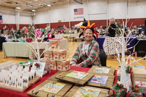 A person with an orange, red, and black hat sits at a table filled with handmade items in a gym with other vendor tables.