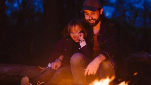 Two people sitting in front of a campfire, wearing coats and laughing together.