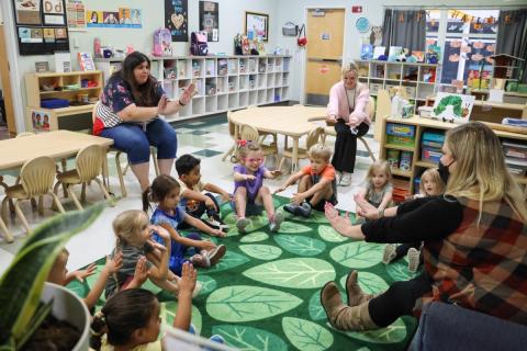 Preschoolers sit in a circle, raising their hands in the air.