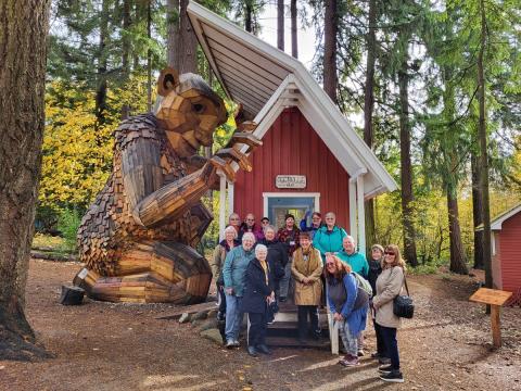 A group of adults standing in front of a small red barn with a wooden troll peeking behind them.