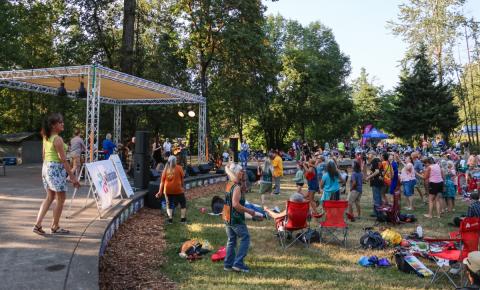 A group of people sit and stand on green grass in front of a large stage with a band playing music.