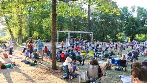 A group of people sit and stand on green grass in front of a large stage with a band playing music.